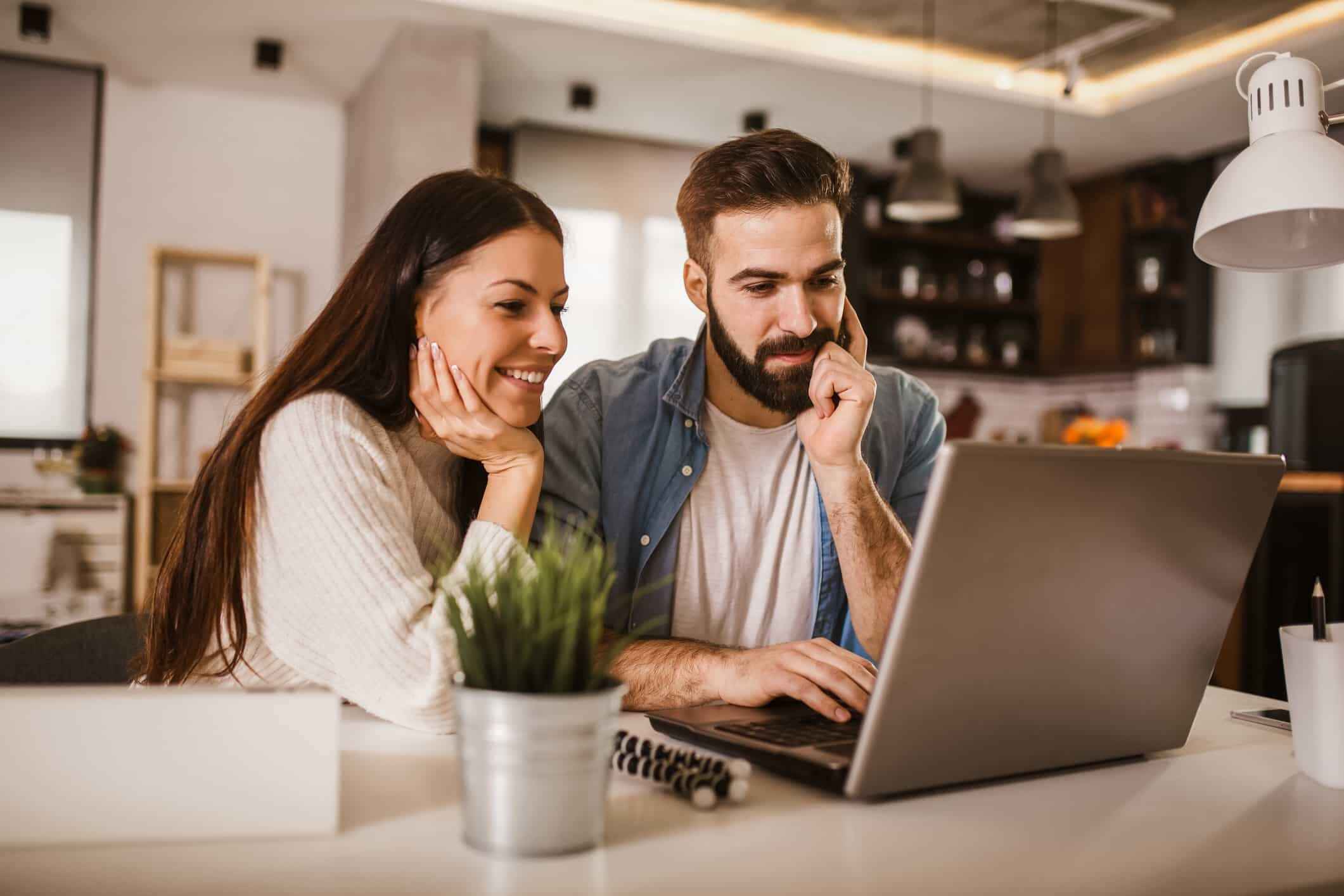 A man and woman, presumably in a romantic relationship, laugh together while looking at something on a laptop.