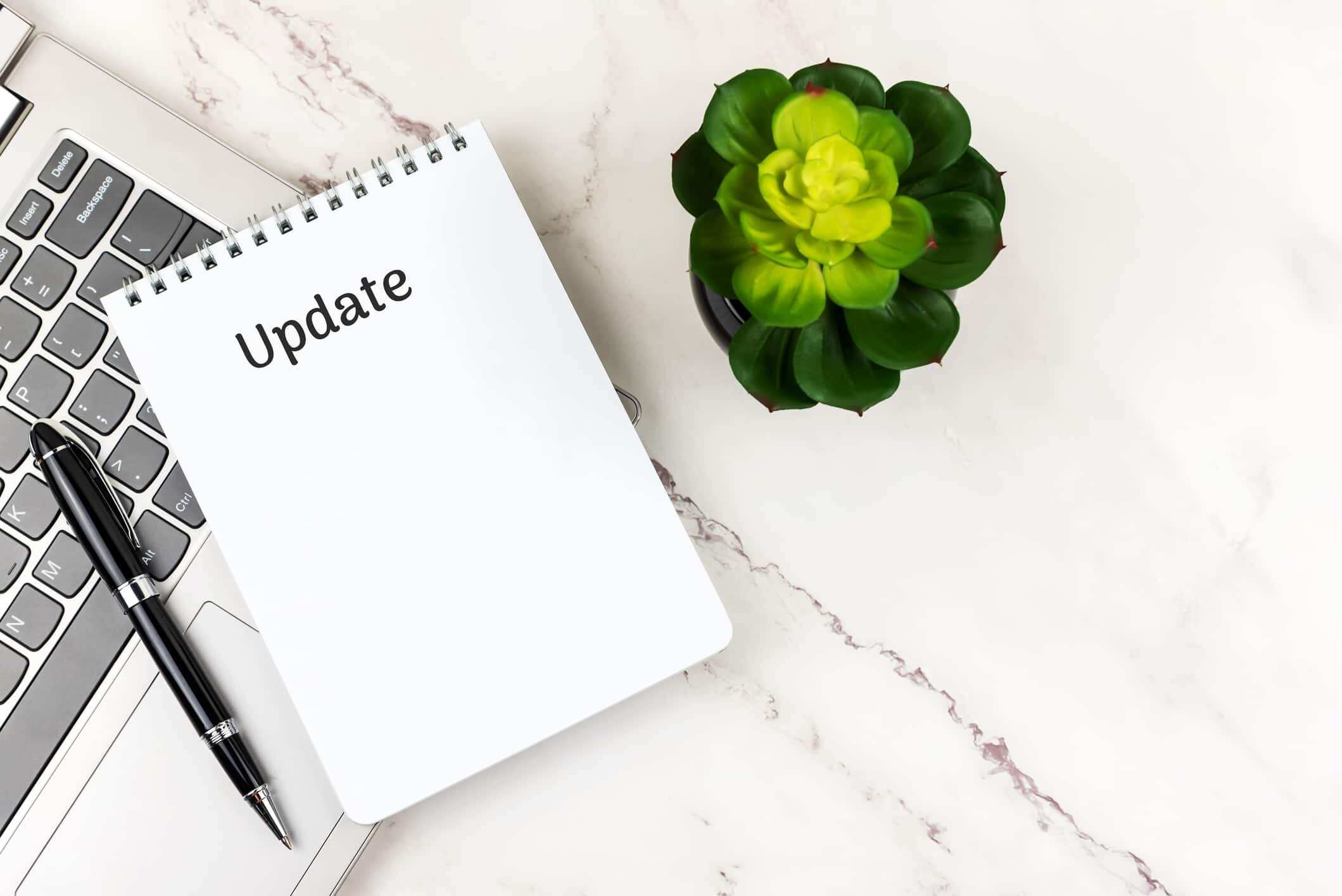 A notepad with the word "Update" written on it sits open on a desk next to a laptop and a potted plant.