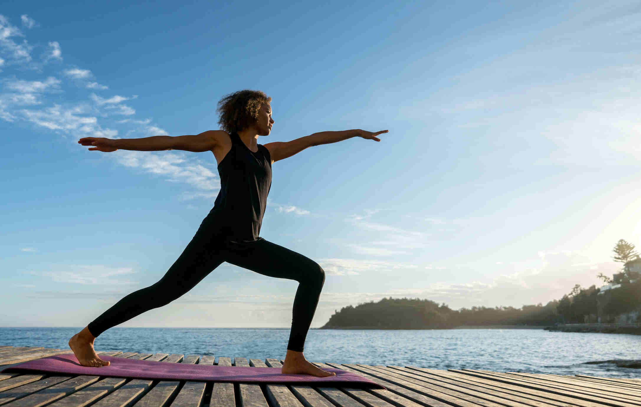 A woman practices yoga stretches outdoors by the sea.