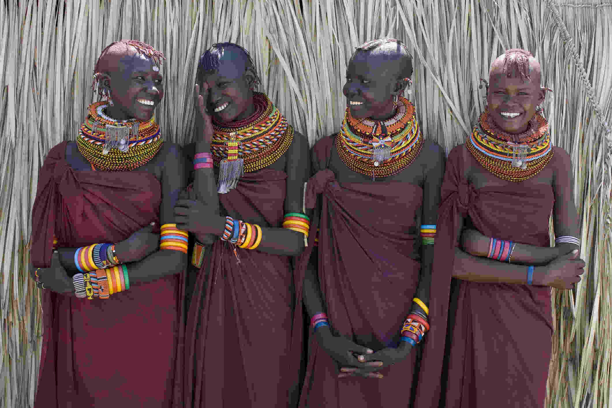 Four young women from the Turkana tribe in Kenya, dressed in traditional attire, stand together smiling.