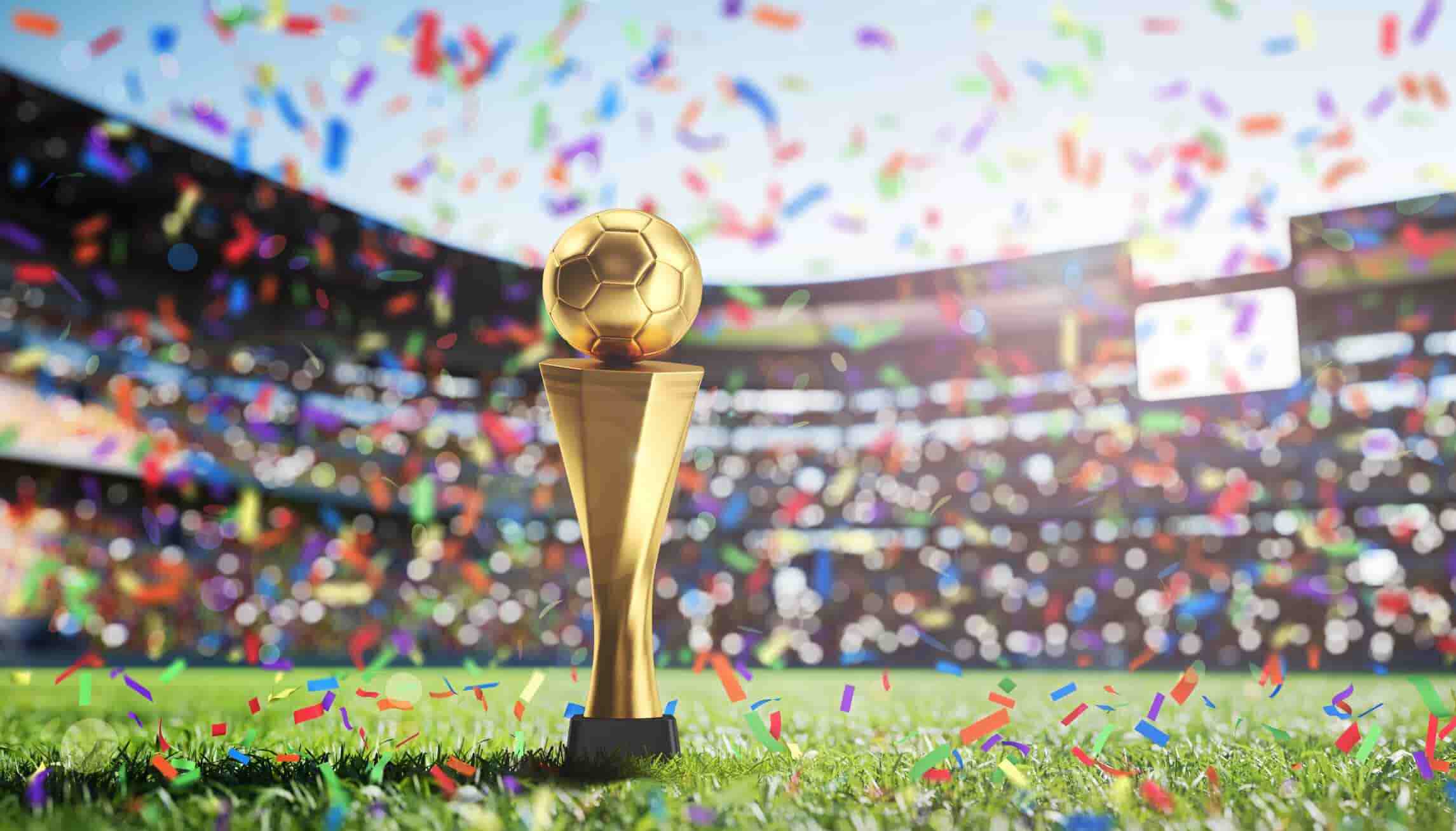 A gold trophy sits on a green field, surrounded by confetti and a blurred background of a cheering crowd in a stadium. The trophy likely represents a championship win in a soccer tournament.