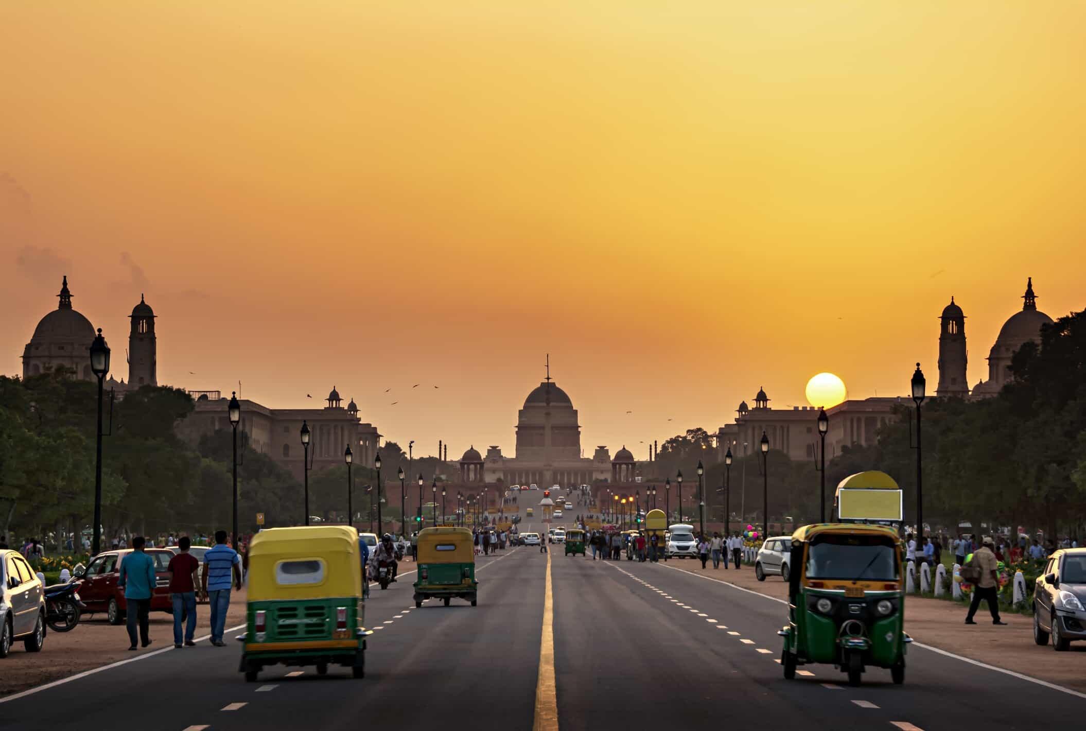 A panoramic view of New Delhi's iconic Rashtrapati Bhavan, illuminated by the setting sun, with traffic and pedestrians filling the wide avenue.