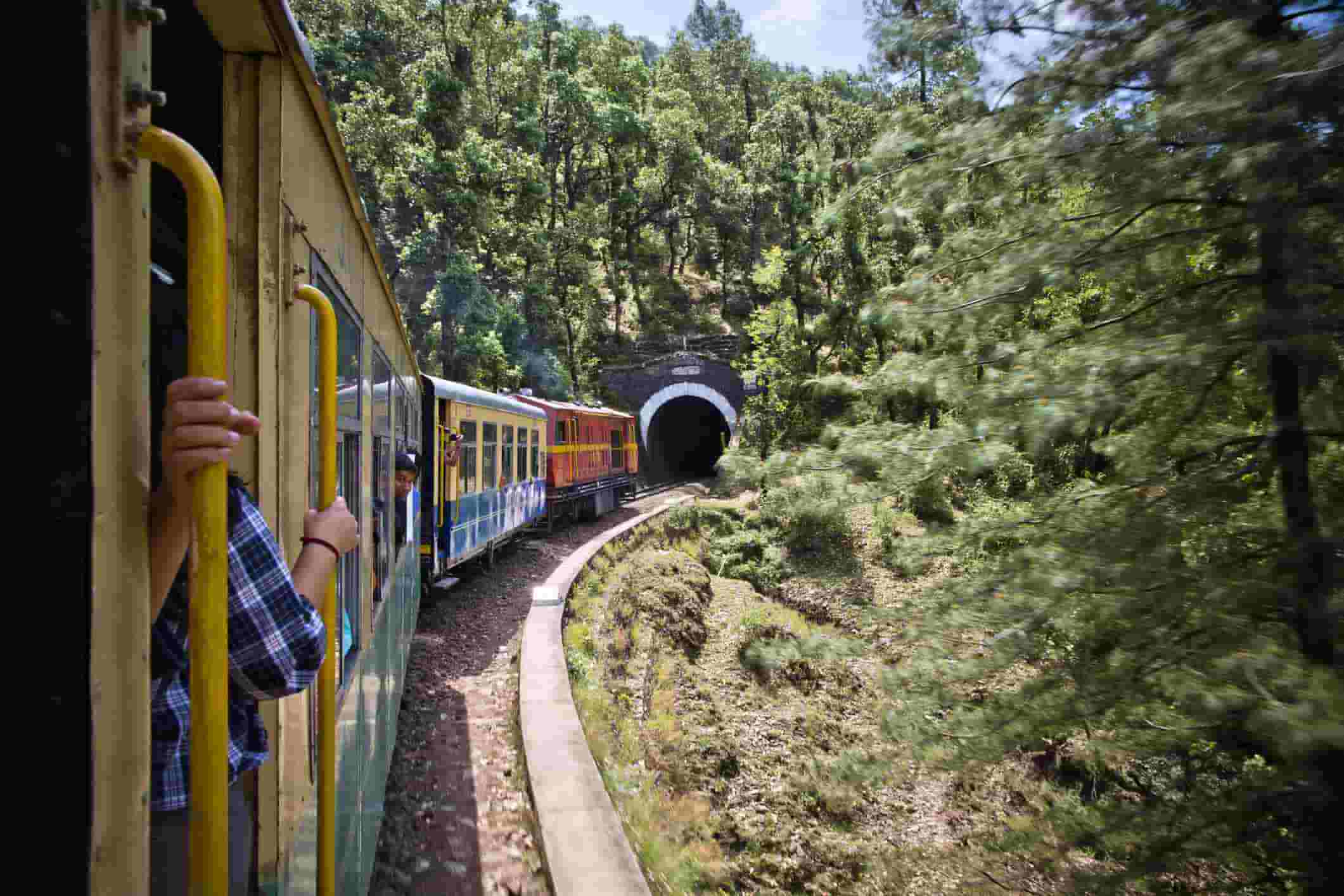 A scenic view from inside a train, showing the train winding through a lush green valley and entering a tunnel.