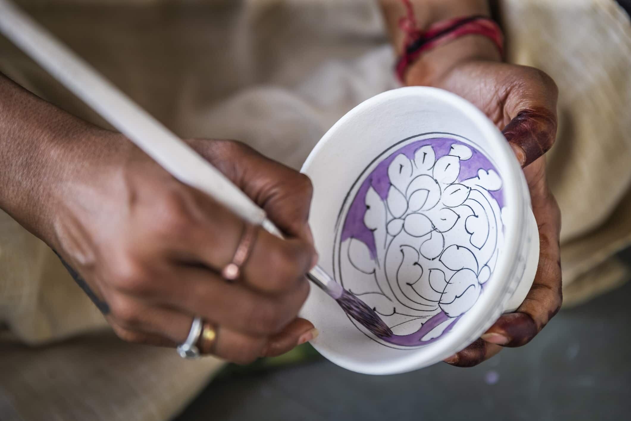 A close-up view of a traditional pottery-making process, featuring a skilled artisan using a brush to create intricate patterns on a blue pottery piece.