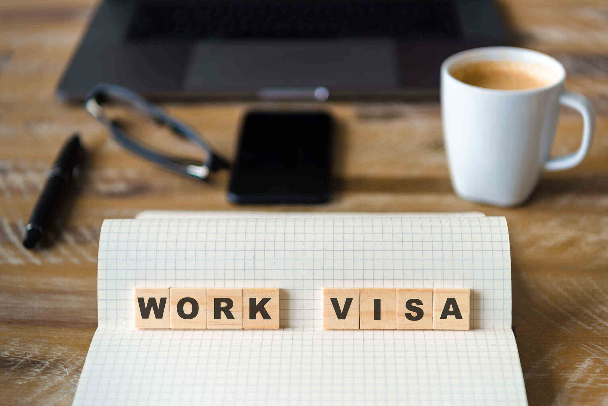 A notebook open on a desk with the words "WORK VISA" arranged in wooden letters. There is a laptop, glasses, a pen, a smartphone, and a coffee cup in the background.