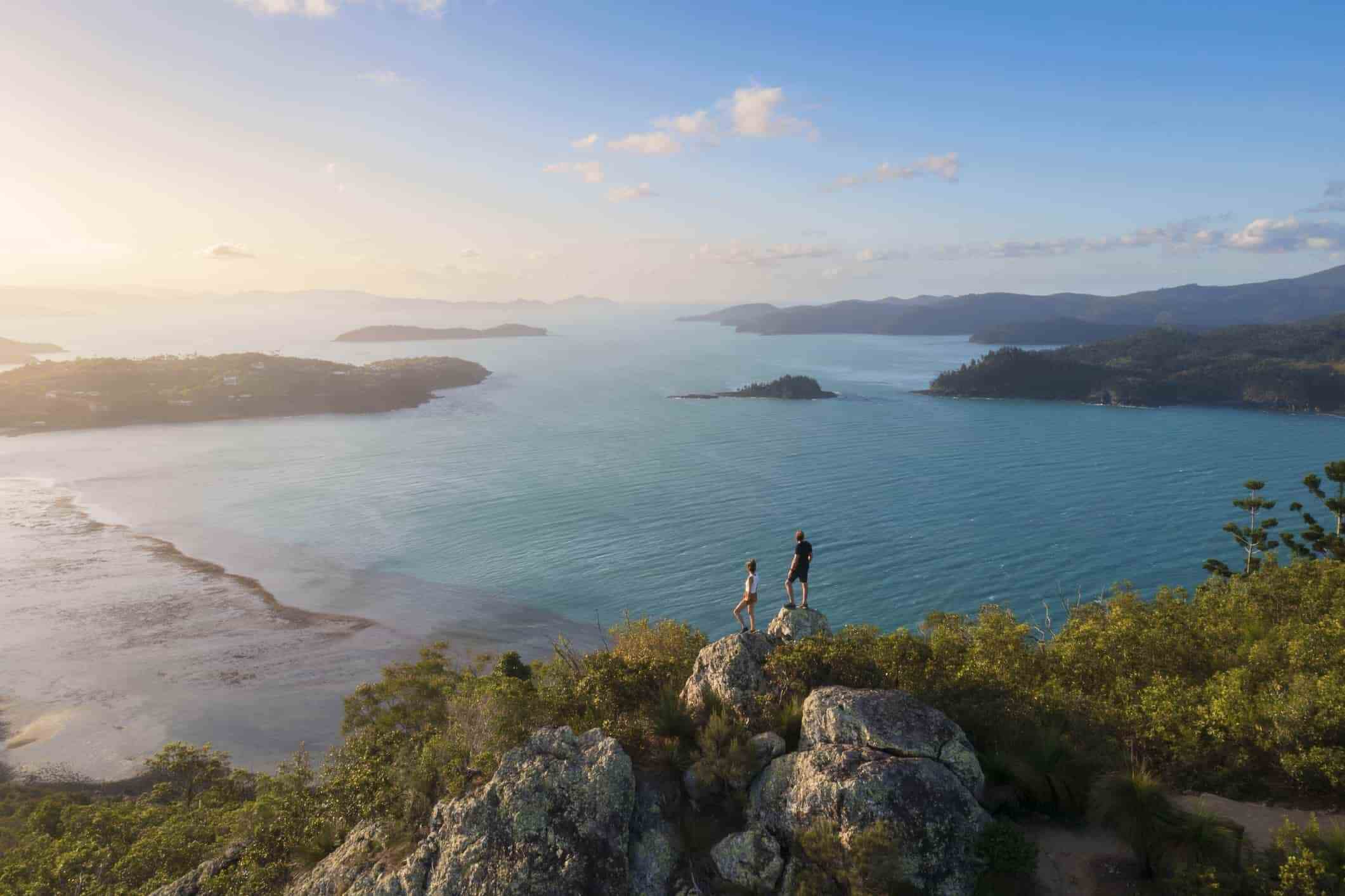 A stunning panorama of a tropical island landscape, featuring lush green hills, sandy beaches, and a vast blue ocean. Two people stand on a cliff, silhouetted against the vibrant sunset.