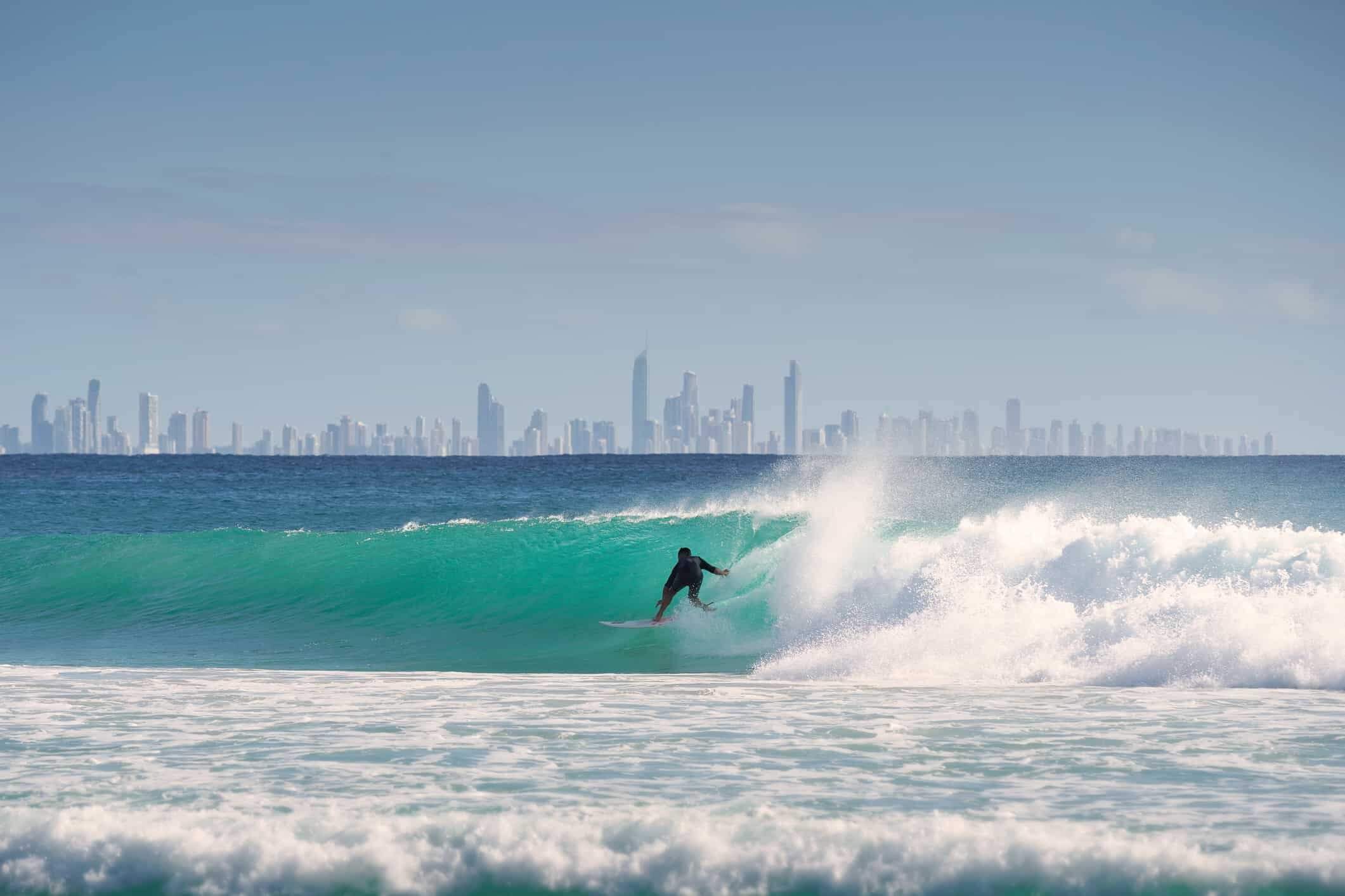 A lone surfer enjoying a large wave, set against the backdrop of a modern coastal city skyline.